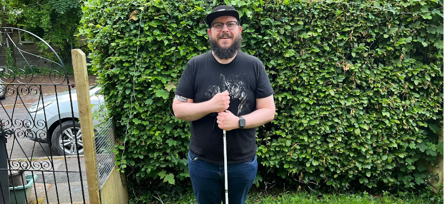 Leigh Holloway standing in front of a green hedge, holding a walking stick and smiling into the camera