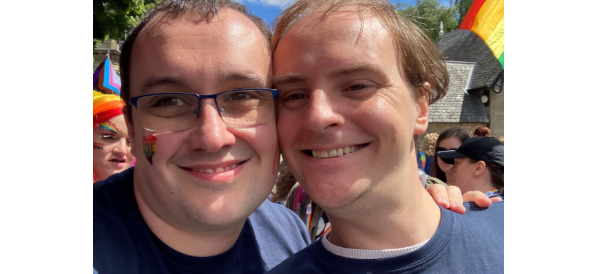 Ross and his husband Tony cheek to cheek smiling. Ross wears glasses and has sticker on his cheek consisting of a rainbow heart with love written within